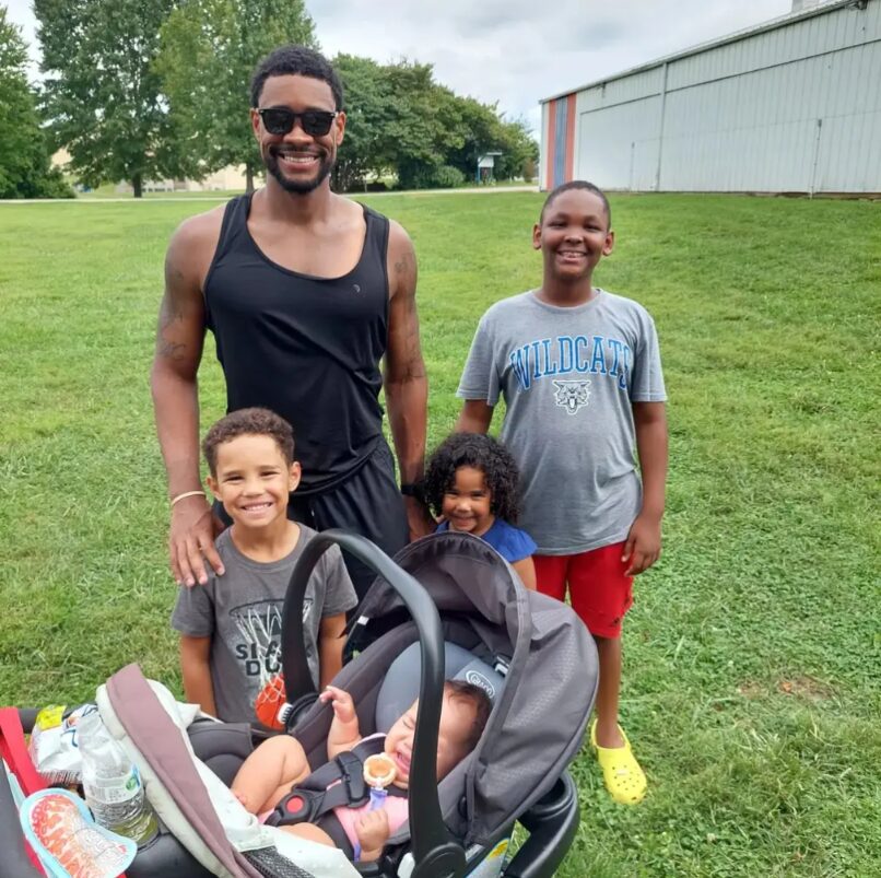 Cameron smiling outdoors on a grassy field with his kids and the baby in a stroller